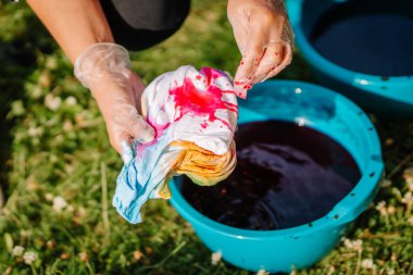 Eldivenli eller, açık hava hippi temalı kravat boyama atölyesi sırasında beyaz gömleğe kırmızı boya sürerler. Renkli kovalar ve parlak renk lekeleri vardır..