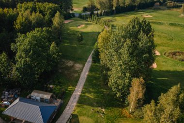 Kum sığınakları, yeşil çim yolları ve çevresi sık ağaçlarla çevrili ve güneş ışığı altında yürüyüş yolu olan bir golf sahasının havadan görünüşü..