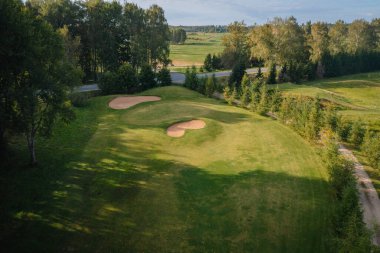 Ağaçlarla çevrili, kırsal bir yol ve parçalı bulutlu bir gökyüzünün altında açık bir kırsal alan olan iki kum sığınağı olan huzurlu bir golf sahası..
