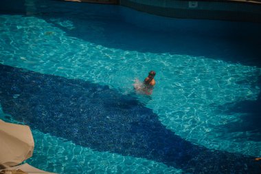 Kemer, Tureky - 18 Eylül 2025 - Güneş ışığı altında açık mavi bir havuzda tek başına yüzen bir adam suya su sıçratıyor. Sahne yukarıdan huzurlu bir yaz anını yakalıyor..