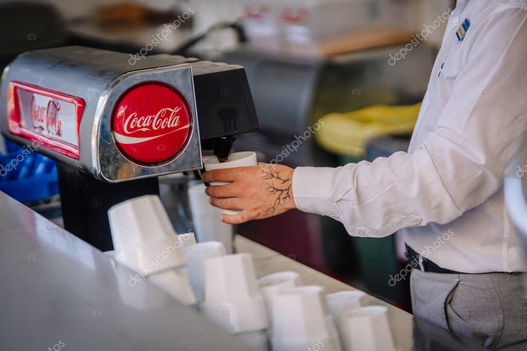 Kemer, Tureky - september 18, 2025 - A staff member in uniform pours a drink from a Coca-Cola soda fountain machine into a cup, with several plastic cups stacked nearby on the counter.