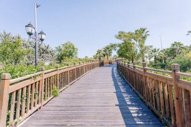Kemer, Tureky - september 18, 2025 - A scenic wooden walkway with railings curves through a lush, green park under a clear blue sky, flanked by palm trees and vintage-style street lamps.