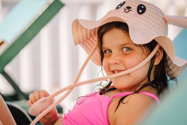Kemer, Tureky - september 18, 2025 - A smiling young girl in a cute animal hat and pink swimsuit relaxes on a lounge chair, enjoying a sunny vacation day, looking playful and happy.