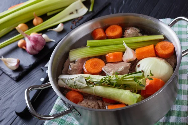 ingredients for veal stew or blanquette de veau - cooked in broth pieces of veal meat in casserole with bouquet garni and vegetables. celery sticks and carrots on cutting board with knife, close-up