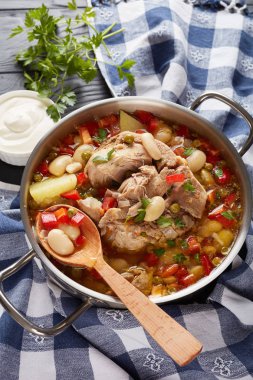 delicious pork vegetable soup with white beans, green peas, red bell pepper, herbs, species in a metal pot with wooden spoon, on a black wooden table, view from above clipart