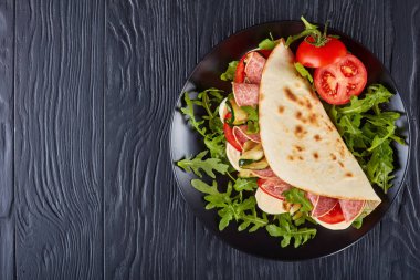 italian piadina with mozzarella, tomato, salami slices, grilled zucchini and arugula on a black plate on a black wooden table, view from above, flat lay clipart