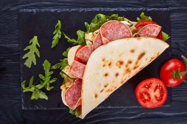 italian piadina with mozzarella, tomato, salami slices, grilled zucchini and arugula on a black slate plate on a wooden table, view from above, flat lay clipart
