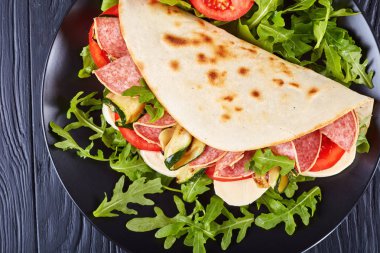 freshly baked italian piadina with mozzarella, tomato, salami slices, grilled zucchini and arugula on a black slate plate on a black wooden table, view from above, close-up, flat lay clipart