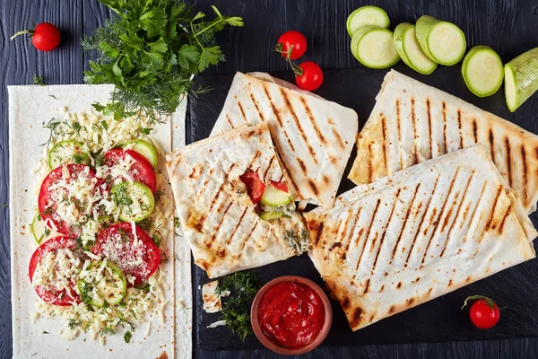 flatbread wraps stuffed with tomato and zucchini slices, greens and grated mozzarella cheese - delicious vegetarian summer lunch served on black slate plate, view from above, flat lay