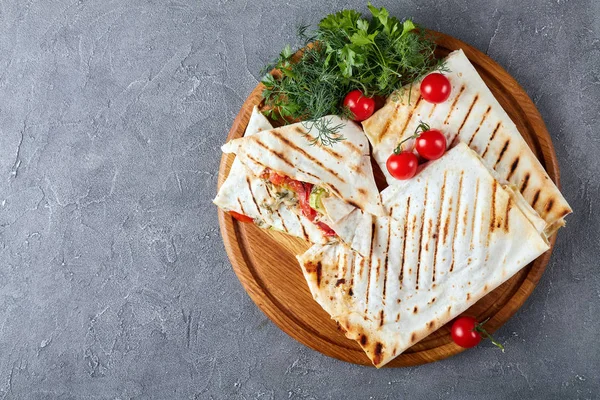 grilled flatbread wraps stuffed with tomato and zucchini slices, greens and grated mozzarella cheese - vegetarian summer lunch on round cutting board on a concrete table, view from above