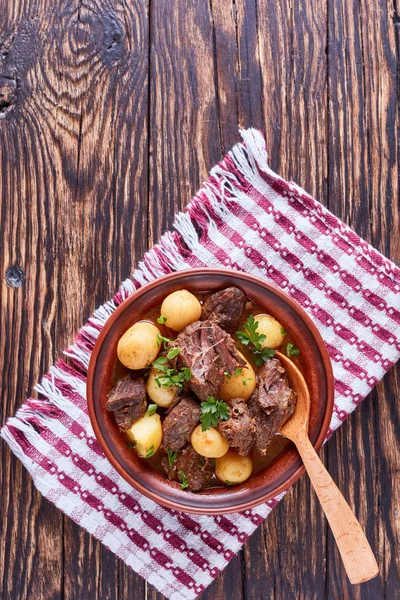 Hot Beef Stew with tender cubes of meat, whole new potatoes, onion and herbs in a clay bowl on old rustic wooden table with napkin, irish cuisine, vertical view from above — Stock Image Hot Beef Stew Tender Cubes Meat Whole New Potatoes Onion Stock Picture