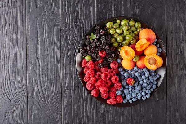 Fruit and berries platter with fresh organic gooseberry, red and black raspberry, blueberry, apricot slices  on a black wooden table, horizontal view from above, Copy space