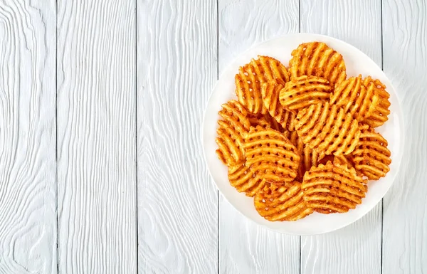 Crispy Potato Waffles Fries, Wavy, Crinkle Cut, Criss Cross Fries on a white plate on a wooden table, view from above, flat lay