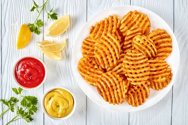 Crispy Potato Waffles Fries, Wavy, Crinkle Cut, Criss Cross Fries on a white plate on a wooden table with mustard and tomato sauce dipping, view from above, flat lay
