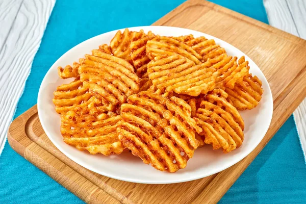 Crispy Potato Waffles Fries, Wavy, Crinkle Cut, Criss Cross Fries on a white plate on a cutting board on a wooden table, view from above, close-up