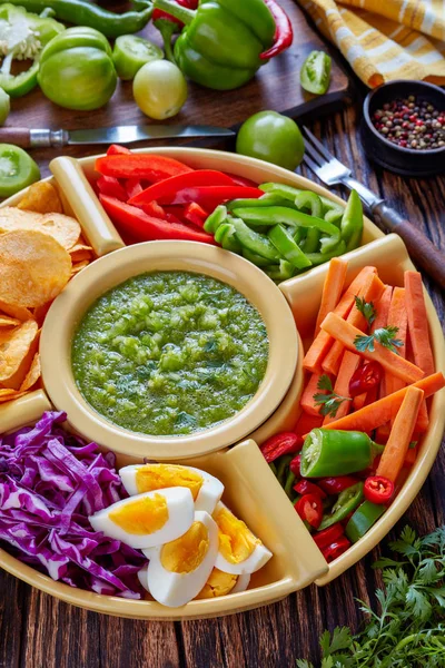 green salsa verde and mexican set of chopped fresh vegetables, hard boiled eggs and chips in bowls on old rustic wooden board with ingredients, vertical view from above