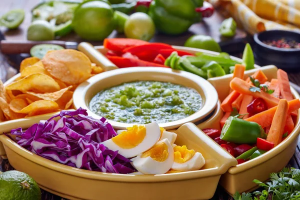 green salsa verde and mexican set for tacos consist of chopped fresh vegetables, hard boiled eggs and chips in bowls on old rustic wooden table with ingredients, horizontal view from above, close-up