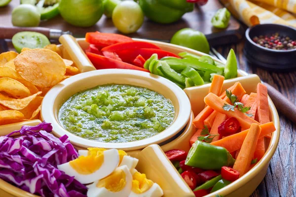 close-up of green salsa verde and mexican set for tacos consist of chopped fresh vegetables, hard boiled eggs and chips in bowls on old rustic wooden table with ingredients, horizontal view from above