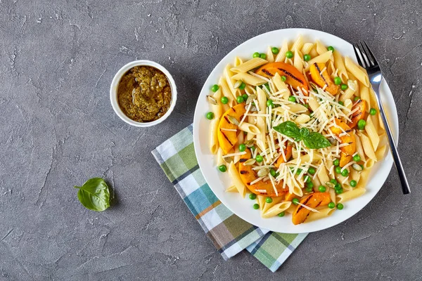 grilled pumpkin and pasta penne with green peas, grated parmesan cheese, and pumpkin seeds on a plate with sauce pesto at the background, view from above, flat lay