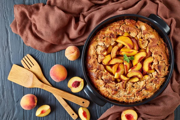 View from above of an old fashioned Peach Cobbler served with fresh ripe peaches, together with kitchen wooden tools: a slotted Turner-Spatula and a flat Turner-Spatula, framed with cotton gauze
