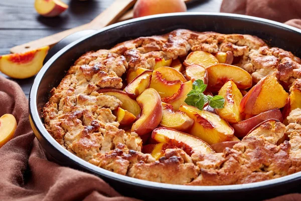 Comfort food dessert: peach crumble in a round black baking dish, close up, with fresh peaches behind the dish