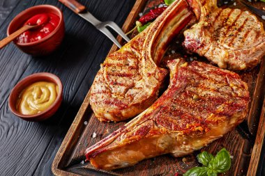 close-up of grilled tomahawk steaks or cowboy beef steaks on an old rude wooden cutting board with fork on a black table with ketchup and mustard, view from above clipart