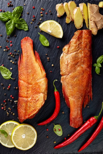 stock image hot smoked red snappers on a black stone plate with spices, herbs and sliced lemons, vertical view from above