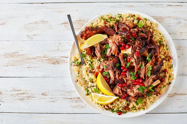 overhead view of couscous parsley salad and slow roast tender leg of lamb pulled off the bones sprinkled with pomegranate seeds on a white platter with spoon, view from above, flat lay, copy space
