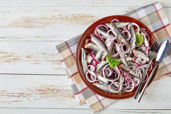 Overhead view of salted sprats marinated with red onion rings, coriander seeds on a plate on a white rustic wooden table with napkin, fish fork and knife, view from above, close-up, flatlay — Stock Image Overhead View Salted Sprats Marinated Red Onion Rings Coriander Seeds Royalty Free Stock Images