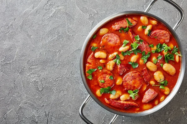 hearty beans stew with sausages, herbs and spices in tomato sauce in a metal casserole on a concrete table, fasolka po bretonsku, polish cuisine, view from above, flatllay, copy space