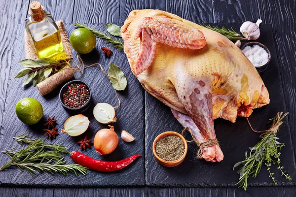 raw whole free-range turkey prepared for marinating on a black stone plate with herbs, lemon, spices on a wooden table, view from above, close-up,