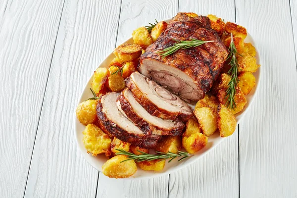 sliced roast pork roulade -  Porchetta, delicious pork roast with crusty roast potatoes served on an oval dish on white wooden table, close-up