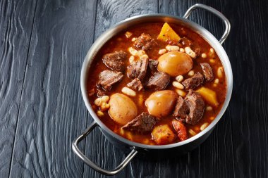 Traditional Jewish Cholent (Hamin) - main dish for the Shabbat lunc, slow cooked beef with potato, beans and brown eggs in a metal casserole on a black wooden table, view from above, close-up clipart