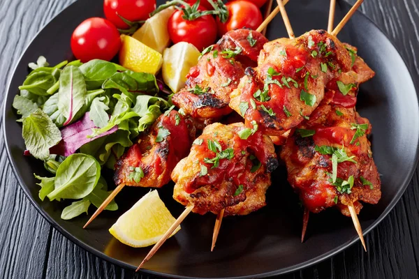 appetizing chicken kebabs chargrilled on skewers served with salad, lemon wedges on a black plate, on a wooden table, view from above, close-up