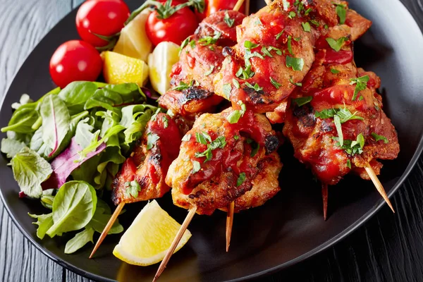 close-up of appetizing chicken kebabs chargrilled on skewers served with salad, lemon wedges on a black plate, on a wooden table, view from above