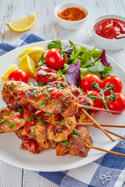 appetizing chicken kebabs chargrilled on skewers served with herbs salad, tomatoes, lemon wedges on a white plate, on a wooden table, view from above