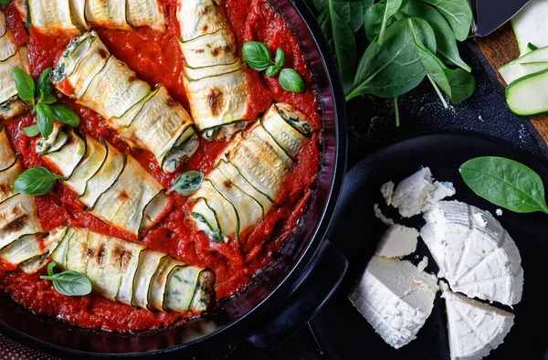 Italian zucchini rolls with cream cheese filling mixed with baby spinach, fresh basil leave baked in tomato sauce served on a black baking dish on a wooden background and fresh ricotta, close-up