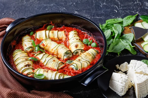 Zucchini strips rolled with a filling of ricotta mixed with baby spinach, fresh basil leave baked in tomato sauce served on a round black baking dish on a  dark concrete background, close-up