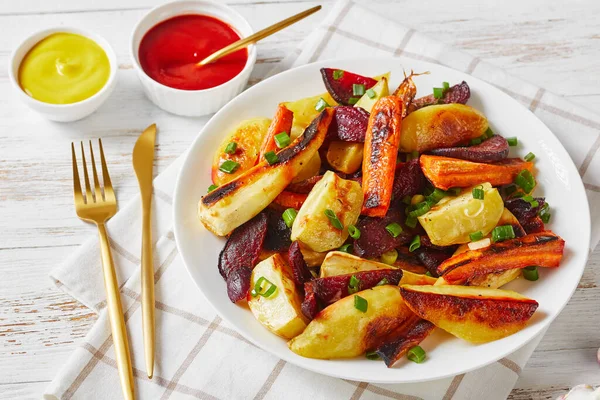 close-up of grilled veggies, beetroot, potato wedges, parsnips, carrots sprinkled with chopped green onion on a white plate with ketchup, mustard, horizontal view from above