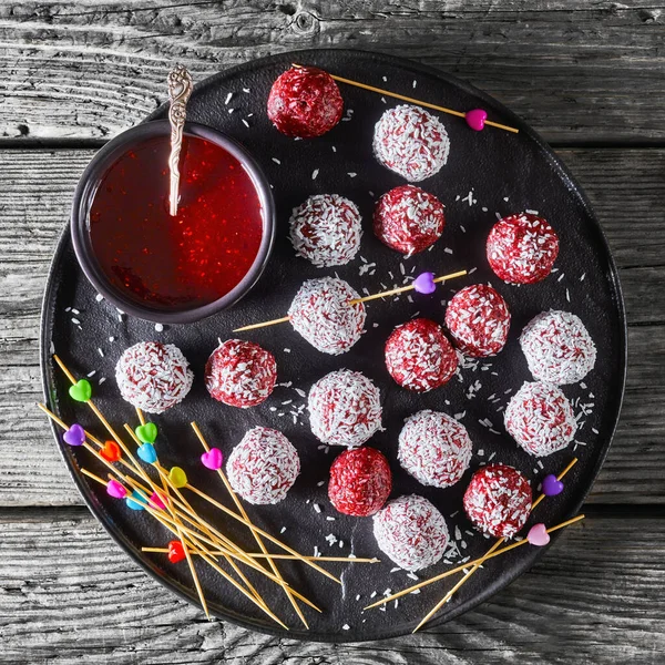 Sugar-free vegetarian dessert: berry bliss balls with desiccated coconut mixed with peanut butter and raspberries, blackberries, blueberries on a black plate on a wooden table, top view, close-up