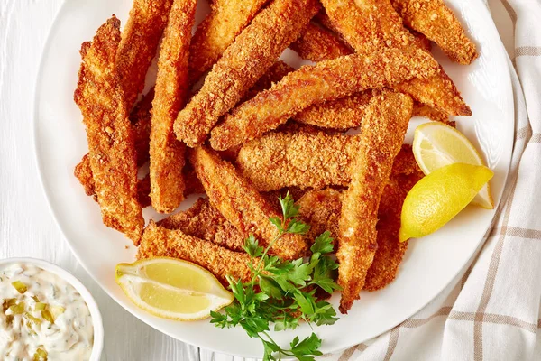 close-up of fish sticks, fish fillet fingers breaded and deep-fried served on a white plate on a wooden table with tartar sauce and lemon wedges, top view, flat lay,