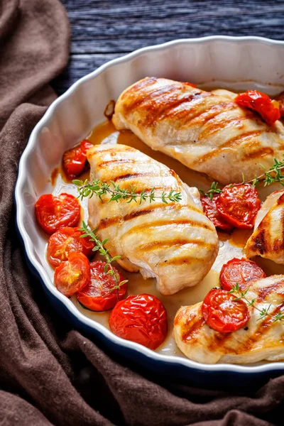 grilled chicken breasts with roasted cherry tomatoes in a baking shell on a dark wooden table with brown cloth, vertical view