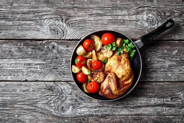 half of roast chicken in a skillet with roasted potato, fresh tomatoes, peppers and herbs on a rustic wooden table, horizontal view from above, close-up, flat lay