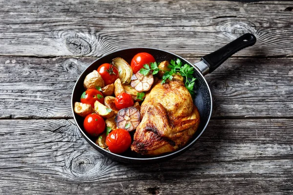 half of roast chicken in a skillet with roasted potato, fresh tomatoes, peppers and herbs on a rustic wooden table, horizontal view from above, close-up