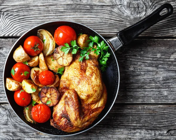 half of roast chicken in a skillet with roasted potato, fresh tomatoes, peppers and herbs on a rustic wooden table, horizontal view from above, close-up, flat lay