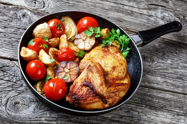 half of roast chicken in a skillet with roasted potato, fresh tomatoes, peppers and herbs on a rustic wooden table, horizontal view from above, close-up