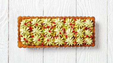 Pistachio ganache tart with pomegranate seeds on a square baking tin made of shortcrust pastry and whipped white chocolate pistachio filling served on a white wooden table, top view, close-up clipart