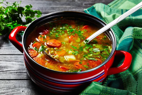 Split pea soup with bavarian sausage, parsnip, carrot topped with parsley and green onion served on a red non-stick saucepan with a soup ladle on a dark wooden background, top view, close-up — Stock Image Split Pea Soup Bavarian Sausage Parsnip Carrot Topped Parsley Green Royalty Free Stock Images