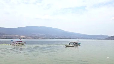 Two boats sailing on a calm lake with mountains in the background and light clouds in the sky, giving a peaceful atmosphere.