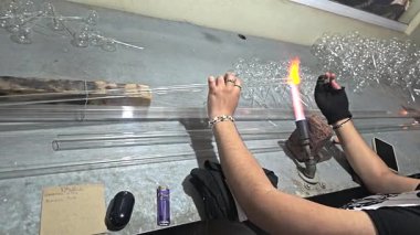 Close-up of a glassblower using flame to shape long glass tubes by hand, with scattered tools and finished pieces nearby.